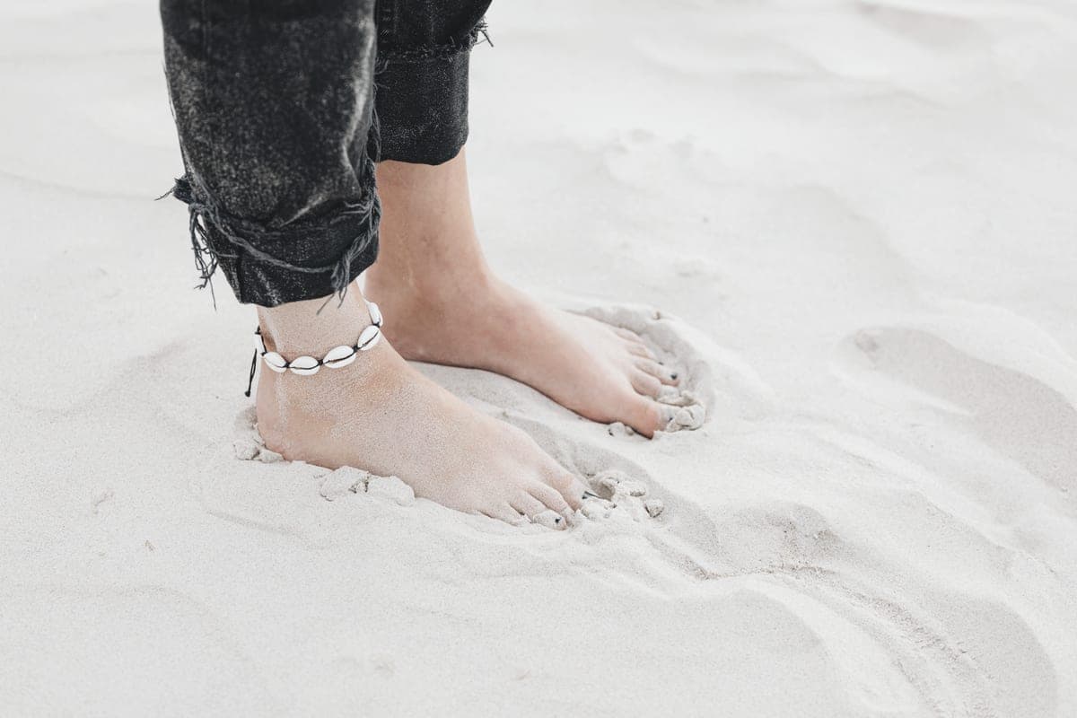 An ankle chain worn on a foot, photographed on sand in daylight