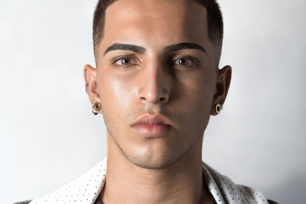 A young man wearing small stud earrings, photographed against a plain pale wall